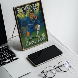 May include: A framed illustration of a Chelsea Football Club player celebrating with a trophy. The artwork is in a comic book style, with the text "CHELSEA FOOTBALL CLUB" and "CHAMPIONS OF EUROPE". The frame is gold-coloured.