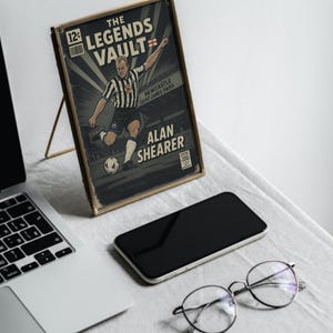 May include: A framed comic-style print featuring Alan Shearer in a Newcastle United uniform kicking a soccer ball. The print is displayed on a gold stand, next to a smartphone, laptop, and round eyeglasses.