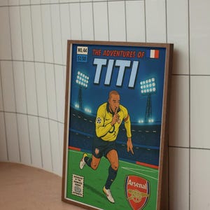 May include: A comic book cover featuring a cartoon illustration of a soccer player in a yellow and blue jersey with the number 3 on the back. The cover is titled "The Adventures of Titi" and features the Arsenal logo. The comic book is framed in a brown wood frame.