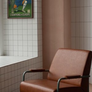 May include: A framed poster of a Chelsea Football Club player with a trophy, text reads "Champions of Europe." The poster is above a brown leather armchair with a metal frame. The room has white tiles and a pink wall.