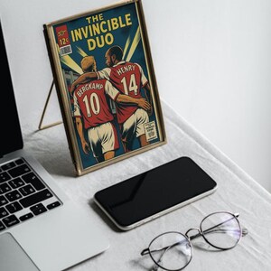 Peut inclure: Une impression encadr&eacute;e de style bande dessin&eacute;e avec le texte "The Invincible Duo", repr&eacute;sentant deux joueurs de football en maillots rouges. L'impression est dans un cadre dor&eacute;, pos&eacute;e sur un support. Un ordinateur portable, un t&eacute;l&eacute;phone et des lunettes sont &eacute;galement visibles.