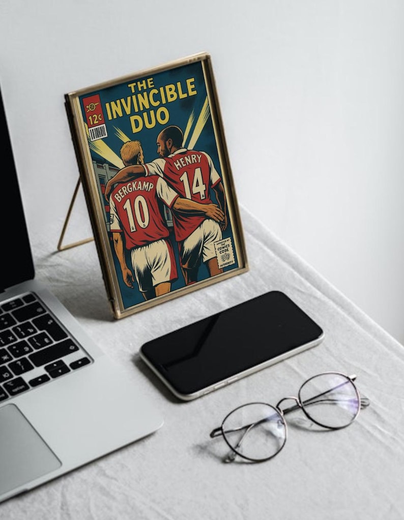 Peut inclure: Une impression encadr&eacute;e de style bande dessin&eacute;e avec le texte "The Invincible Duo", repr&eacute;sentant deux joueurs de football en maillots rouges. L'impression est dans un cadre dor&eacute;, pos&eacute;e sur un support. Un ordinateur portable, un t&eacute;l&eacute;phone et des lunettes sont &eacute;galement visibles.