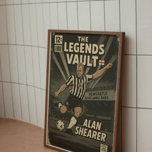 May include: A framed vintage-style print featuring a soccer player kicking a ball. The print reads "The Legends Vault" and "Alan Shearer" with the Newcastle Saint James Park stadium in the background. The color palette includes black, white, and brown.