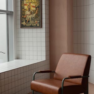 Puede incluir: Un sill&oacute;n de cuero marr&oacute;n con patas y reposabrazos de metal. Un p&oacute;ster enmarcado en la pared presenta una ilustraci&oacute;n de estilo c&oacute;mic de un jugador de f&uacute;tbol con el texto "THE GOLDEN KANE". La habitaci&oacute;n tiene baldosas blancas y una pared rosa.