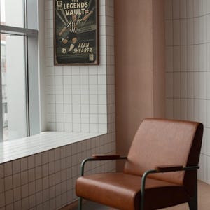 May include: A framed vintage poster titled "The Legends Vault" featuring Alan Shearer in a soccer action pose. A brown leather armchair with metal legs sits in front of a tiled wall and window.