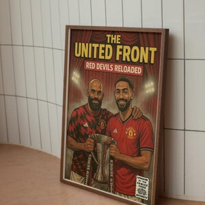 May include: Framed poster featuring two men in red soccer jerseys holding a trophy. The poster has the text "THE UNITED FRONT RED DEVILS RELOADED" in yellow and red. The image is set against a white tiled wall.