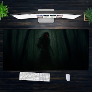 May include: A large rectangular desk mat featuring a dark forest scene with a shadowy figure with glowing red eyes. A white keyboard, mouse, and computer monitor are visible on a dark wood surface.