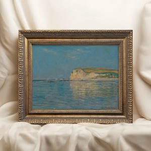 May include: A framed landscape painting depicting a coastal scene with a cliff, beach, and ocean. The artwork features a gold ornate frame. The painting uses blue, green, and tan hues to depict the sky, water, and land. The artwork is displayed on a white cloth.