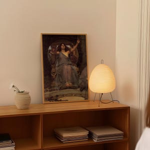 May include: A wooden framed print of a classical painting depicting a woman in a flowing gown. A white paper lantern lamp with black legs sits on a wooden shelf, next to a small beige vase with a white flower.