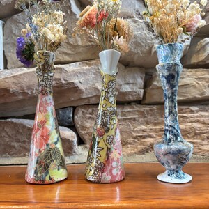 May include: Three slender bud vases holding dried flowers. The left vase has a pink and floral pattern, the center vase features a yellow and floral design, and the right vase displays a blue and white marbled pattern with a flared base.