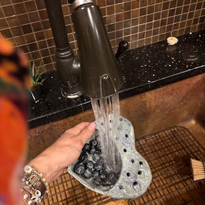 May include: A heart-shaped ceramic berry colander with a speckled blue and grey glaze, featuring multiple drainage holes, being held under a kitchen faucet while water rinses a cluster of fresh blueberries inside the bowl.