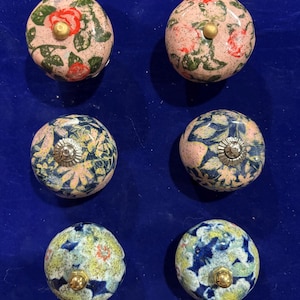 Peut inclure: Six boutons de placard ronds en céramique disposés en deux colonnes. La paire supérieure présente une base rose avec des motifs floraux rouges et une quincaillerie dorée. La paire centrale a une base bleu foncé avec des motifs floraux clairs et une quincaillerie argentée. La paire inférieure affiche un motif floral tacheté bleu et jaune avec une quincaillerie dorée.