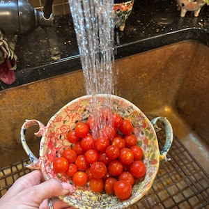 May include: A ceramic colander with two handles and a colorful floral pattern filled with bright red cherry tomatoes being rinsed under a stream of water from a kitchen faucet. The bowl features numerous drainage holes throughout its surface.