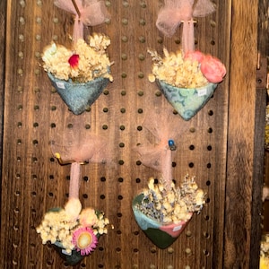 May include: Four heart-shaped ceramic wall planters in shades of blue, green, and pink, each containing dried floral arrangements. Each planter is suspended by a light pink tulle ribbon tied in a bow, hanging from a dark brown wooden pegboard.