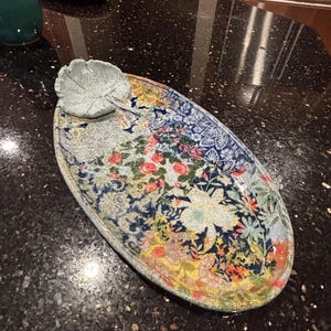 Puede incluir: Una bandeja de servicio ovalada de cerámica con un vibrante patrón floral multicolor en tonos azul, amarillo, rojo y verde. Un pequeño plato texturizado en forma de hoja está unido a la parte superior izquierda de la superficie.