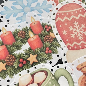 Pu&ograve; includere: Una collezione di etichette di carta fustellate con illustrazioni natalizie che includono un fiocco di neve azzurro, una muffola rossa con motivo a fiocco di neve, una ghirlanda verde con candele rosse e una tazza verde con marshmallow.
