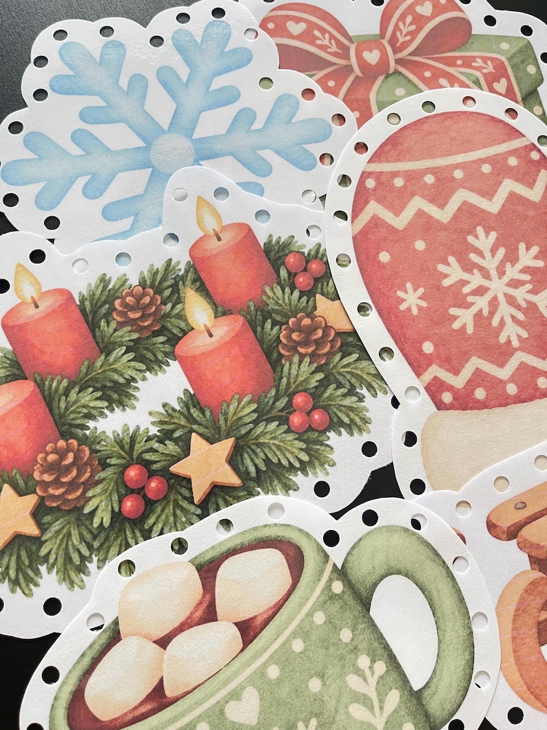 Pu&ograve; includere: Una collezione di etichette di carta fustellate con illustrazioni natalizie che includono un fiocco di neve azzurro, una muffola rossa con motivo a fiocco di neve, una ghirlanda verde con candele rosse e una tazza verde con marshmallow.