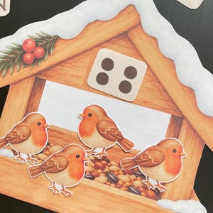 Pu&ograve; includere: Un'illustrazione fustellata di una mangiatoia per uccelli in legno coperta di neve, con quattro piccoli pettirossi marroni e arancioni appollaiati sul bordo. La mangiatoia contiene un mix di semi ed &egrave; decorata con un rametto di bacche rosse. &Egrave; visibile una tessera quadrata con quattro punti.