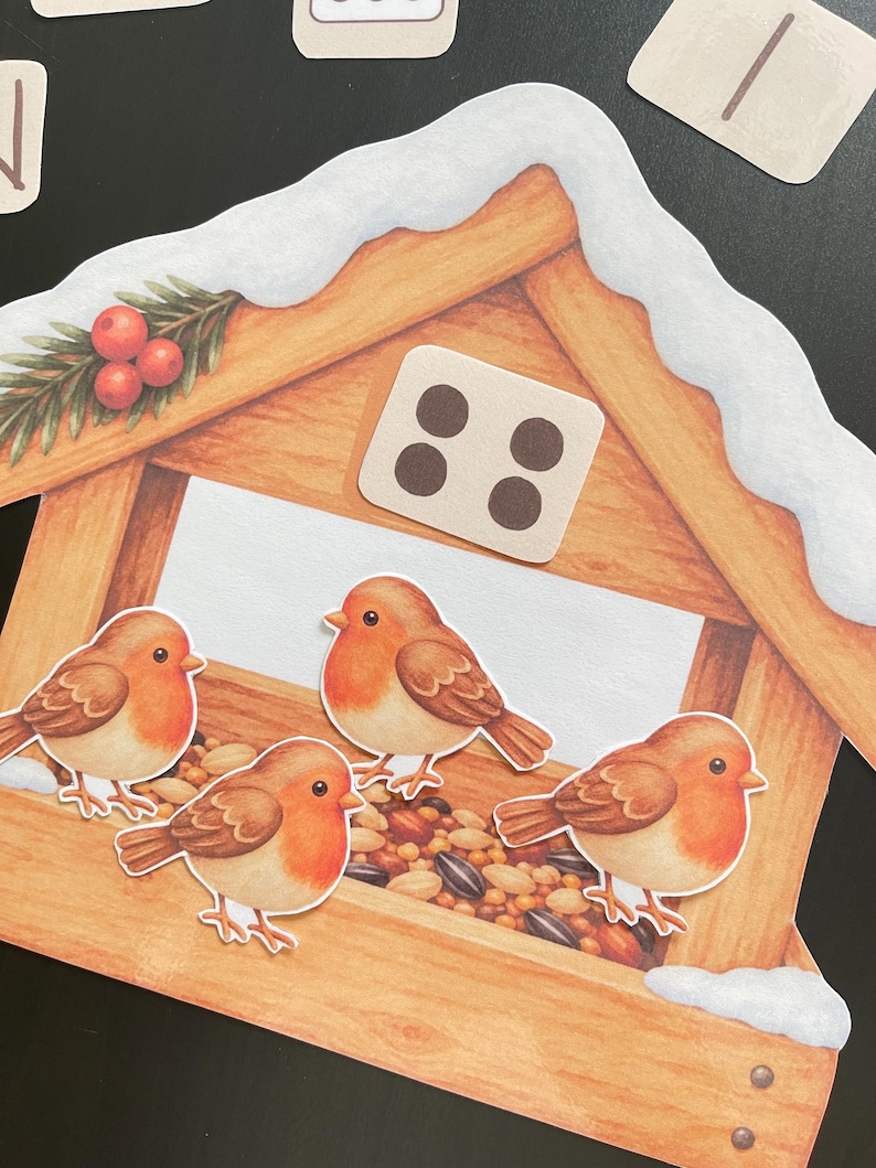 Pu&ograve; includere: Un'illustrazione fustellata di una mangiatoia per uccelli in legno coperta di neve, con quattro piccoli pettirossi marroni e arancioni appollaiati sul bordo. La mangiatoia contiene un mix di semi ed &egrave; decorata con un rametto di bacche rosse. &Egrave; visibile una tessera quadrata con quattro punti.