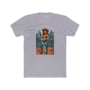 May include: A heather gray t-shirt featuring a cartoon illustration of a cowgirl holding a gun. The cowgirl is wearing a yellow top, blue shorts, and boots. Text on the shirt reads "Sexy and From Texas."