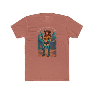 May include: A terracotta-colored t-shirt featuring a graphic of a cowgirl holding a gun in front of a saloon. The text "Sexy and From Texas" is above the graphic. The shirt is made of a soft, comfortable material.