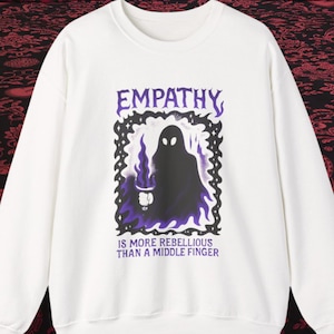 May include: White sweatshirt with a graphic design. The design features the word "EMPATHY" in purple above a black silhouette holding a torch, framed by a purple border. The text "IS MORE REBELLIOUS THAN A MIDDLE FINGER" is below the graphic.