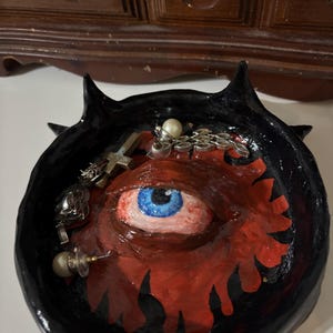 Peut inclure: Un vide-poches rond noir fait à la main avec un motif d'œil rouge et noir. Le plat a de petites cornes et contient des bijoux, notamment des boucles d'oreilles, un pendentif en forme de croix et une chaîne. L'œil a un iris bleu.