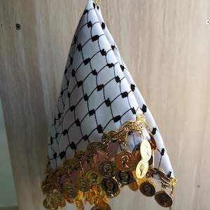 May include: A white and black keffiyeh scarf featuring a repeating diamond grid pattern. The bottom edge is adorned with a decorative gold-colored trim and a series of dangling gold-toned circular coins, all suspended from a gold metal ring.