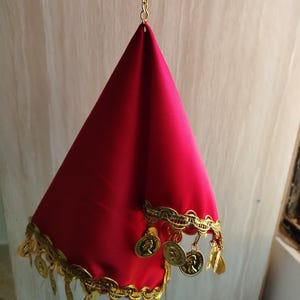 May include: A vibrant red fabric cone suspended from a gold-toned metal ring by a short chain. The base of the cone is adorned with a decorative gold-colored trim featuring dangling metallic coin-shaped charms that hang along the bottom edge.