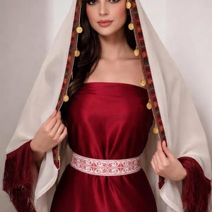 May include: A deep red strapless dress paired with a white head covering featuring a dark patterned border, gold coin accents, and long red fringe at the ends. A white belt with a detailed red geometric pattern is worn around the waist.