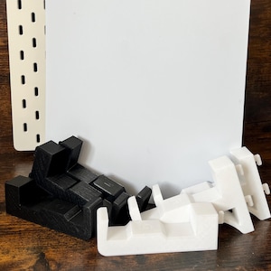 Könnte beinhalten: Eine weiße PlayStation 5-Konsole mit dem PlayStation-Logo oben links. Die Konsole wird von schwarzen und weißen Zubehörteilen begleitet, wahrscheinlich zur Unterstützung oder Aufbewahrung. Der Hintergrund ist eine Holzoberfläche.