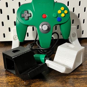 Könnte beinhalten: Grüner Nintendo 64-Controller mit schwarzem Kabel und einem Satz schwarzer und weißer Controller-Ständer. Der Controller hat eine graue Mitte, eine rote A-Taste und gelbe, blaue und grüne Tasten.