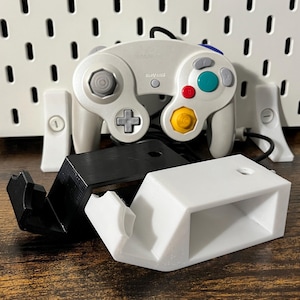 Könnte beinhalten: Ein weiß-grauer Videospiel-Controller mit bunten Knöpfen, einem schwarzen Kabel und zwei weißen und schwarzen Controller-Ständern. Der Controller liegt auf einer Holzoberfläche.