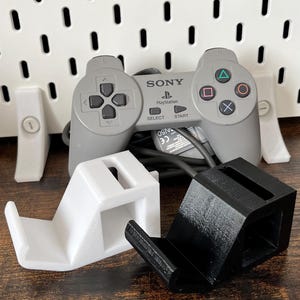 Könnte beinhalten: Ein grauer PlayStation-Controller mit schwarzem Kabel und zwei Controller-Ständern, einer weiß und einer schwarz. Der Controller hat das Sony-Logo und das PlayStation-Logo. Die Ständer sind zur Aufnahme des Controllers konzipiert.