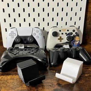 Könnte beinhalten: Eine Auswahl an Videospiel-Controllern, darunter ein weiß-schwarzer PlayStation 5-Controller, ein weiß-goldener Controller im Zelda-Design und zwei schwarze Controller. Außerdem zwei Controller-Ständer, einer schwarz und einer weiß.