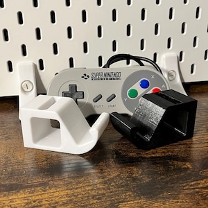 Könnte beinhalten: Ein Super Nintendo Controller mit zwei 3D-gedruckten Controller-Haltern, einer weißen und einer schwarzen. Der Controller ist grau mit farbigen Knöpfen und dem Text "SUPER NINTENDO". Die Halter sind zur Montage des Controllers konzipiert.
