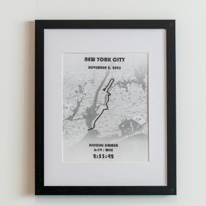 May include: Framed black and white map print of New York City, with a route highlighted in black. The text "NEW YORK CITY NOVEMBER 2, 2025" is at the top. Below the map are the words "NICOLAS SIMMER 6:39 / MILE 2:53:42".