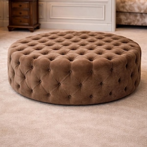 May include: A large, circular ottoman in a warm brown colour. The ottoman has a tufted design with evenly spaced button detailing. The plush, upholstered piece sits on a neutral-coloured carpet, suggesting a living room or lounge setting.