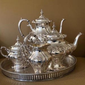 Könnte beinhalten: Ein silbernes Teeservice, bestehend aus einer Teekanne, einem Sahnekännchen, einer Zuckerdose und einem Serviertablett. Das Set hat aufwendige Details und wird auf einem runden Tablett präsentiert. Das Teeservice ist aus glänzendem Silbermetall gefertigt.