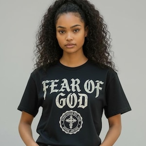 May include: Black t-shirt with the text "FEAR OF GOD" in a distressed font. Below the text is a circular emblem with a cross. The model is wearing light blue denim shorts.