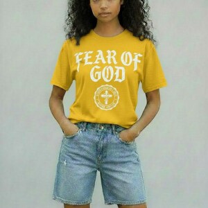 May include: Mustard yellow t-shirt with the text "FEAR OF GOD" in white, along with a circular emblem. The shirt is paired with light blue denim shorts. The model is standing with hands in pockets.