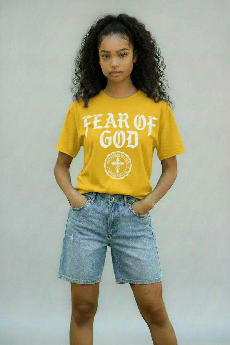 May include: Mustard yellow t-shirt with the text "FEAR OF GOD" in white, along with a circular emblem. The shirt is paired with light blue denim shorts. The model is standing with hands in pockets.