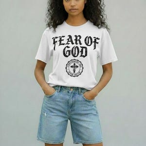 May include: White t-shirt with the text "FEAR OF GOD" in a bold, gothic font. Below the text is a circular emblem with a cross and the words "STREETWEAR EST. 2012 DIVINE JUSTICE". The model is wearing light blue denim shorts.
