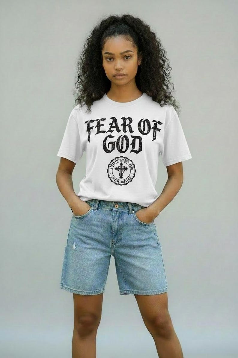 May include: White t-shirt with the text "FEAR OF GOD" in a bold, gothic font. Below the text is a circular emblem with a cross and the words "STREETWEAR EST. 2012 DIVINE JUSTICE". The model is wearing light blue denim shorts.