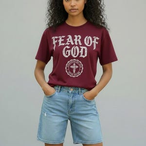 May include: A maroon t-shirt with the text "FEAR OF GOD" in a distressed white font, along with a circular emblem. The model is wearing light blue denim shorts. The outfit is casual and stylish.