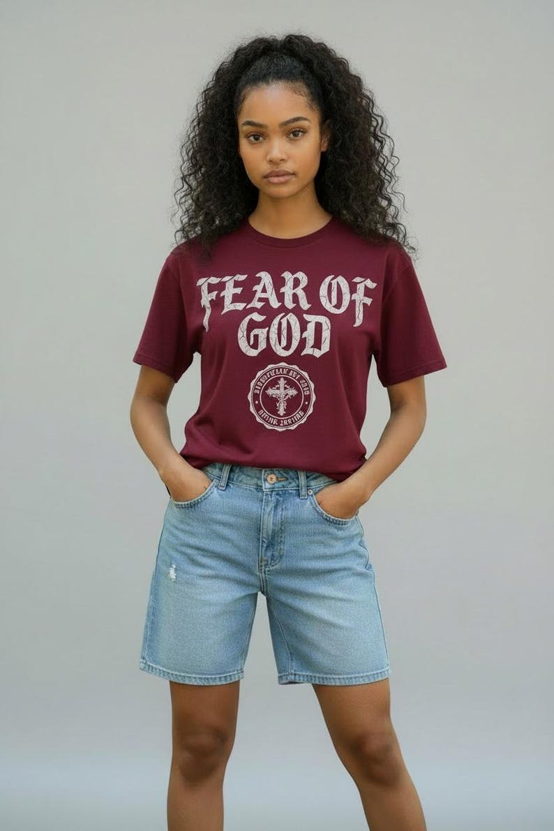 May include: A maroon t-shirt with the text "FEAR OF GOD" in a distressed white font, along with a circular emblem. The model is wearing light blue denim shorts. The outfit is casual and stylish.