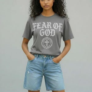 May include: A gray t-shirt with the text "FEAR OF GOD" in a distressed white font, paired with light blue denim shorts. The shirt features a circular graphic with a cross. The model is wearing the outfit.