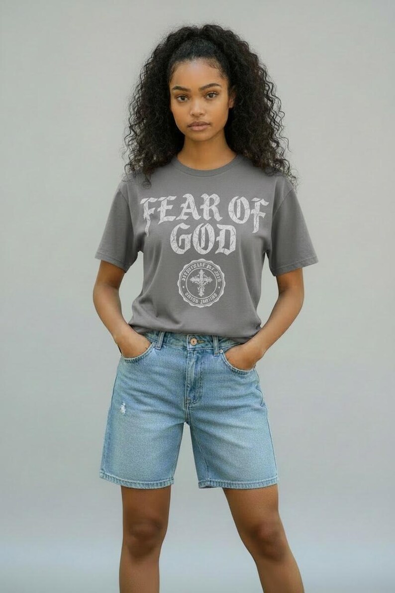 May include: A gray t-shirt with the text "FEAR OF GOD" in a distressed white font, paired with light blue denim shorts. The shirt features a circular graphic with a cross. The model is wearing the outfit.