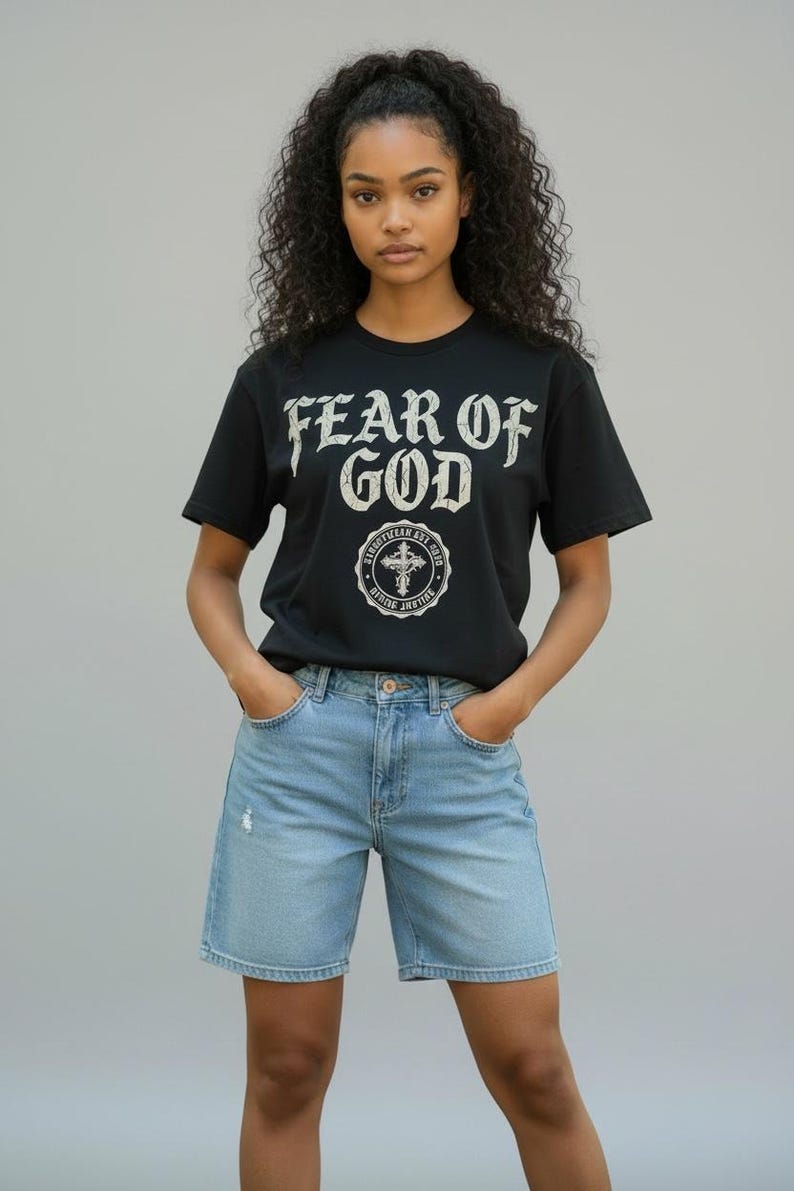 May include: Black t-shirt with the text "FEAR OF GOD" in a distressed font. Below the text is a circular emblem with a cross. The model is wearing light blue denim shorts.
