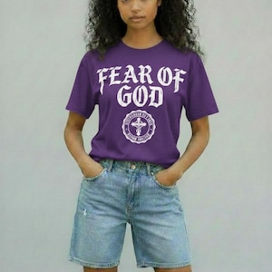 May include: A purple t-shirt with the text "FEAR OF GOD" in white, above a circular emblem with a cross. The model is wearing light blue denim shorts. The background is a solid light gray.